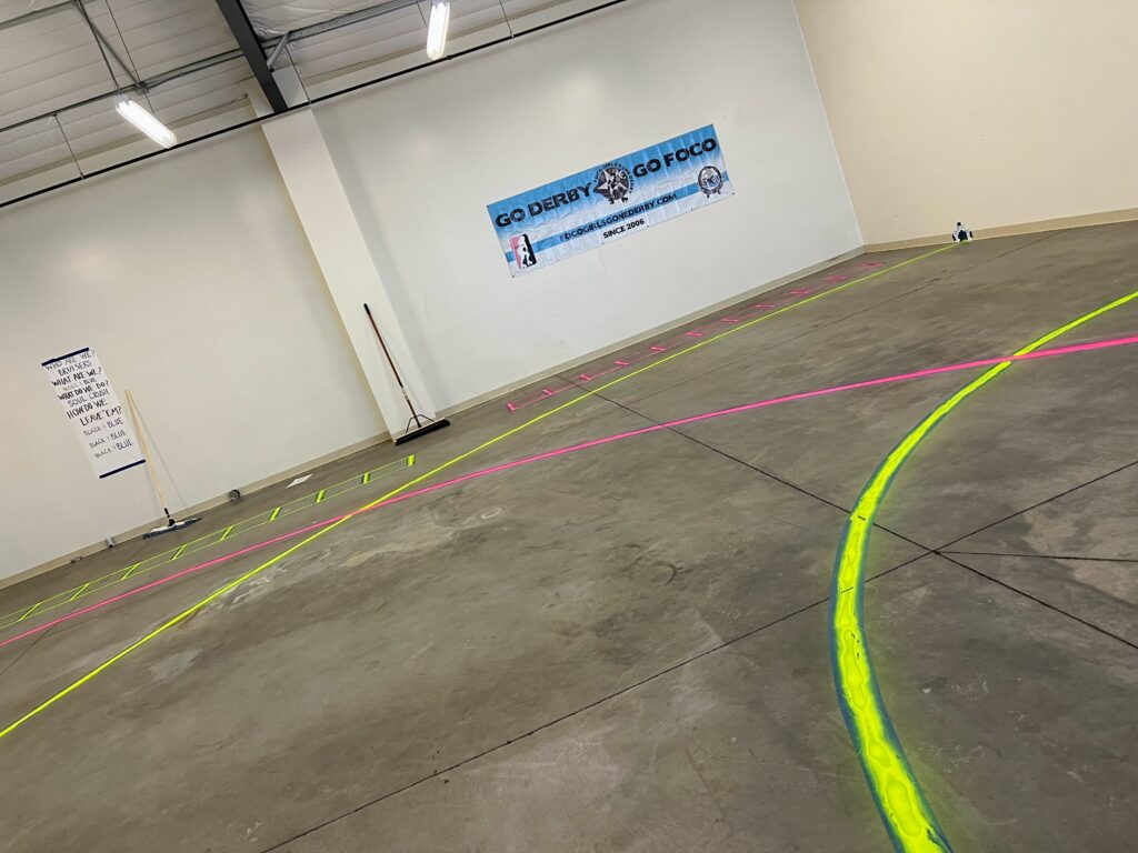 Inside a warehouse with concrete floors. Overlapping quarters of roller derby track are painted on the floor in hot pink and hot yellow. There are FoCo Roller Derby posters on the walls.