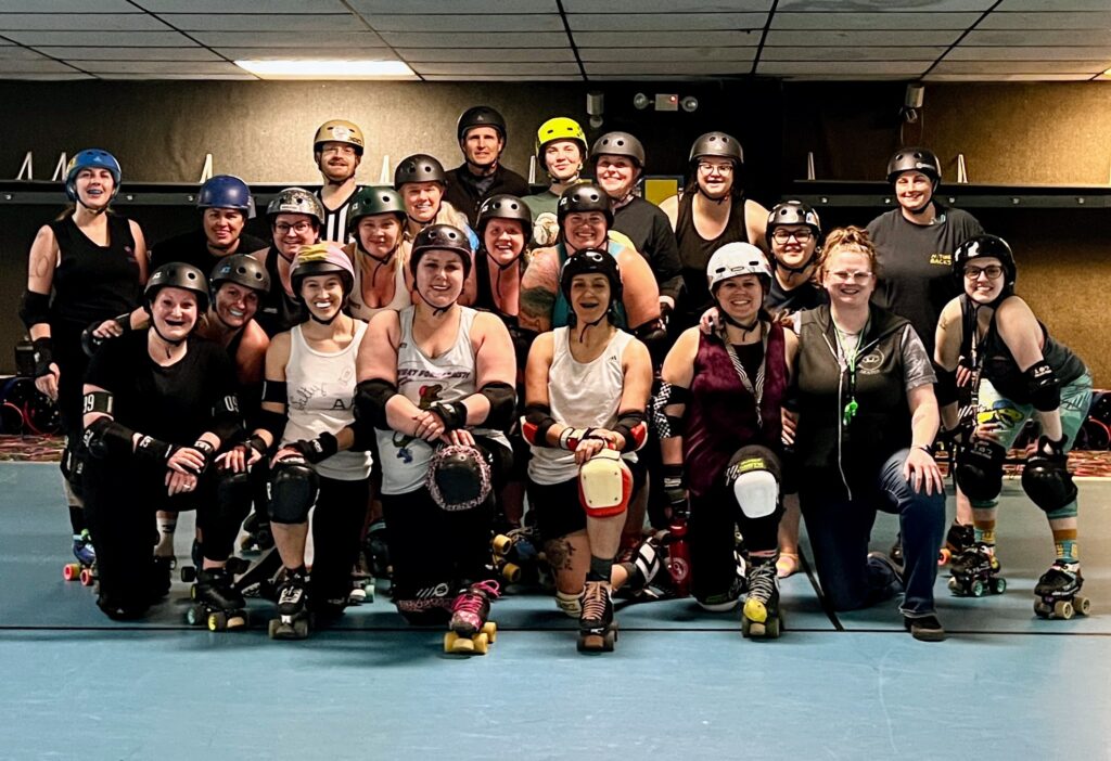 A group of roller derby skaters and officials pose in three rows after scrimmage.