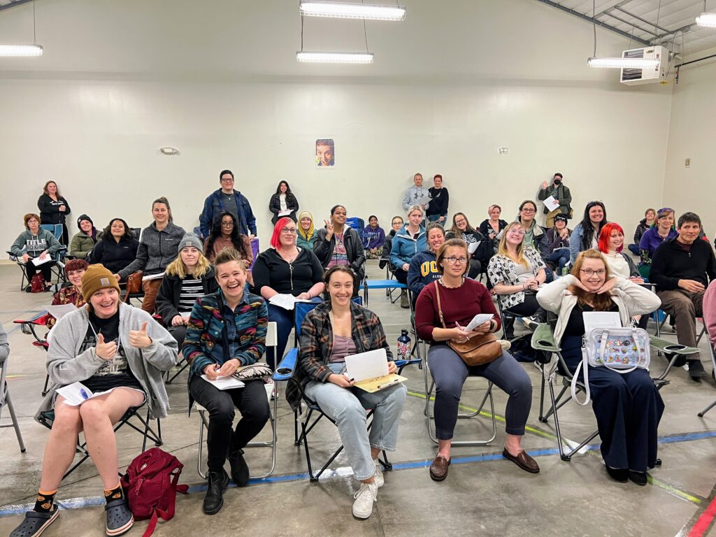 A group of people sitting in chairs in a warehouse look at the camera. Some are smiling and posing.